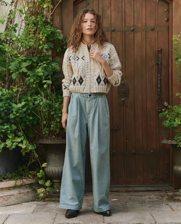 A woman stands before a wooden door, surrounded by green plants, wearing a patterned cream sweater, black shoes, and The Sawyer Pant in Boiler Wash by THE GREAT. PF25—wide-leg light blue jeans.