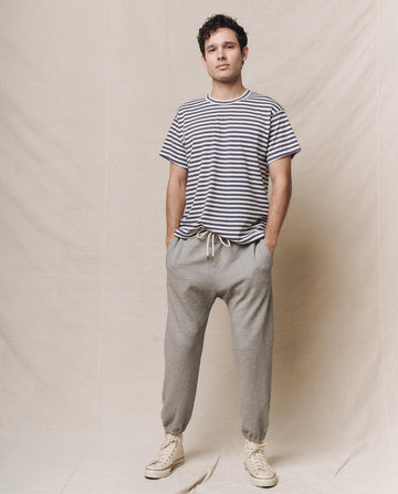 Standing against a beige backdrop, the person wears The Men's Boxy Crew in Vintage Blue Stripe by THE GREAT. SP25 POLO LOUNGE MEN, paired with gray joggers and cream sneakers, hands casually tucked in pockets.