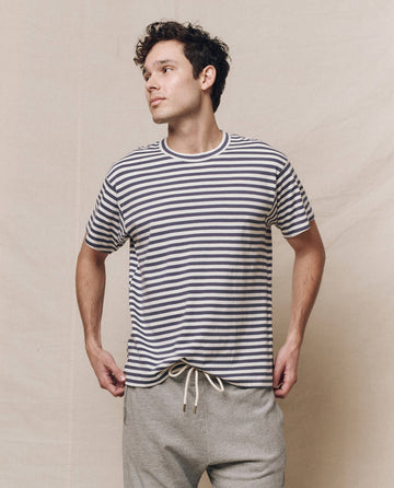 A person wearing The Men's Boxy Crew in Vintage Blue Stripe by THE GREAT. SP25 Polo Lounge Men pairs it with gray pants, standing against a plain backdrop, looking to the side with hands in pockets.