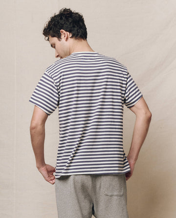 A person in a Men's Boxy Crew from THE GREAT. SP25 POLO LOUNGE MEN, featuring vintage blue stripes, and gray pants stands with their back to the viewer against a plain background.
