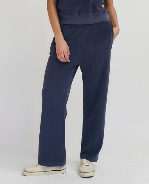 A person stands against a plain background wearing THE GREAT. SU25 MICRO TERRY The Micro Terry Victory Sweatpant in Vintage Blue with a matching top and white sneakers, one hand casually in their pocket.