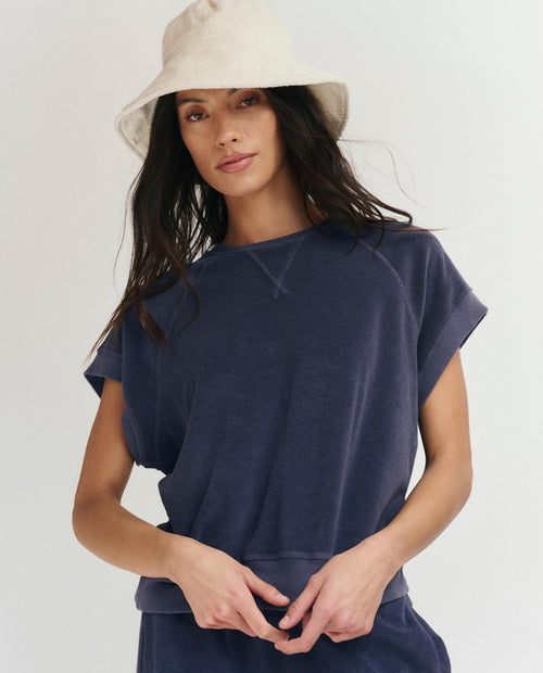 A woman wearing THE GREAT.'s The Micro Terry Wedge Sweatshirt in Vintage Blue stands against a plain background, looking at the camera.