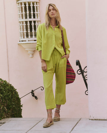 A woman stands outdoors against a pale pink wall, wearing loose-fitting Citron The Reef Pant by THE GREAT. SP25 SWIM with a matching shirt, flat woven shoes, and a striped shoulder bag.