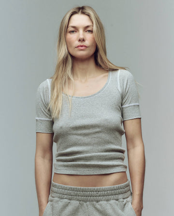 A woman with long blonde hair wears THE GREAT. SU25 BABY RIB The Baby Rib Ballet Tee in Heather Grey, styled with grey sweatpants, and stands against a plain background.