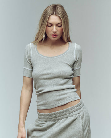 A woman with long blonde hair wears THE GREAT. SU25 BABY RIB The Baby Rib Ballet Tee in Heather Grey, paired with grey sweatpants, standing against a plain light background and looking down.