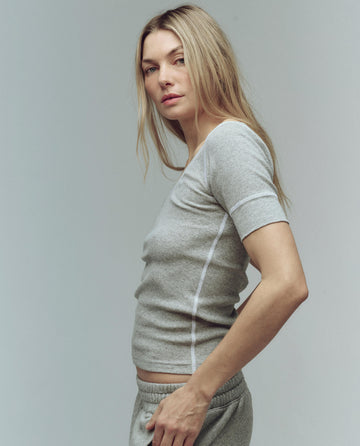 A woman with long blonde hair stands sideways against a plain background, wearing THE GREAT. SU25 BABY RIB The Baby Rib Ballet Tee in Heather Grey with matching grey pants.