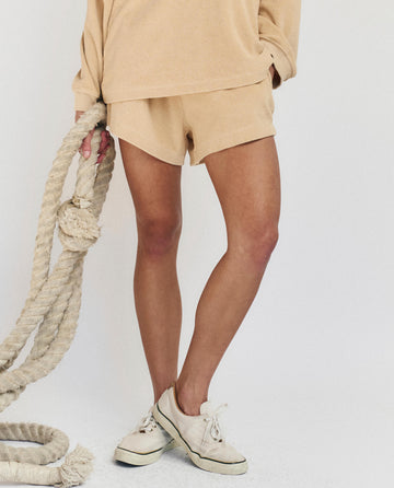 Wearing THE GREAT. SU25 MICRO TERRY The Micro Terry Swing Short in Chamois and a beige microterry sweatshirt, this person pairs the set with white sneakers, holding a thick rope against a plain light backdrop.