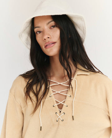 Model wearing THE GREAT. SU25 MICRO TERRY Micro Terry Lace Up Tunic in Chamois with a white bucket hat, looking at the camera against a plain background.