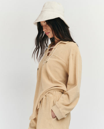 A woman in THE GREAT. SU25 MICRO TERRY The Micro Terry Lace Up Tunic in Chamois with matching pants and a light bucket hat stands against a plain background, looking down.