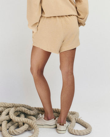 A person stands with their back to the camera, wearing THE GREAT. SU25 MICRO TERRY "The Micro Terry Swing Short" in Chamois with a matching top and white sneakers, beside coiled thick rope on a white floor.