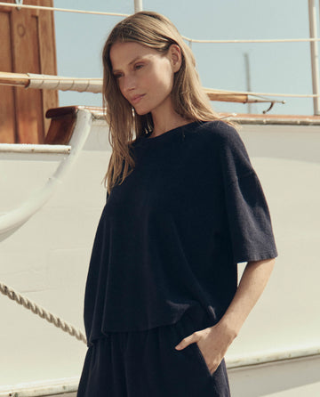 A woman with long hair stands outdoors near a boat, wearing THE GREAT. SU26’s The Micro Terry Track Crew in Navy, one hand in her pocket and looking slightly downward.