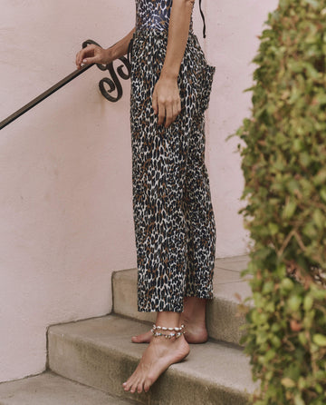 A person wears THE GREAT. SP25 SWIM's The Reef Pant – Leopard, stands barefoot on outdoor steps near a pink wall and green bush, holding a metal handrail.