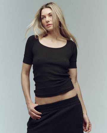 A woman with long blonde hair wears THE GREAT. SU25 BABY RIB The Baby Rib Ballet Tee in Almost Black, paired with black pants, standing against a plain light background.