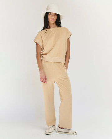 A woman stands against a plain background wearing THE GREAT. SU25 MICRO TERRY The Micro Terry Wedge Sweatshirt in Chamois with matching pants, a white bucket hat, and white sneakers.