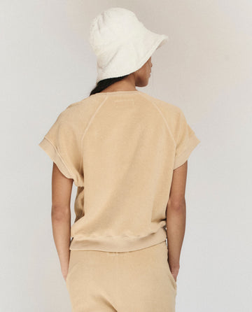 A person wears THE GREAT. SU25 MICRO TERRY Wedge Sweatshirt in Chamois, matching pants, and a white bucket hat, standing with their back to the camera against a neutral background.