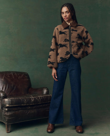 A person with braids stands beside a worn brown leather chair, wearing THE GREAT. F25 D2's The Pasture Jacket in Light Hickory, wide-leg blue jeans, and brown shoes, set against a green backdrop.
