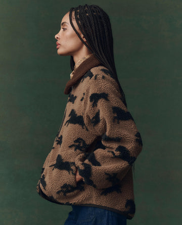 A person with long braids poses in profile, wearing THE GREAT. F25 D2 The Pasture Jacket in Light Hickory and blue jeans, set against a green background.