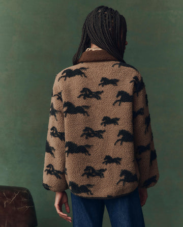 Seen from behind, a person with braided hair wears THE GREAT. F25 D2's The Pasture Jacket in Light Hickory, featuring a black horse pattern, along with blue jeans, standing before a green background.