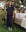 A person wearing THE GREAT. PS25 SLEEP's The Short Sleeve Pajama Shirt in Mini Black Polka Dot stands by a bed outdoors, surrounded by trees and bushes.