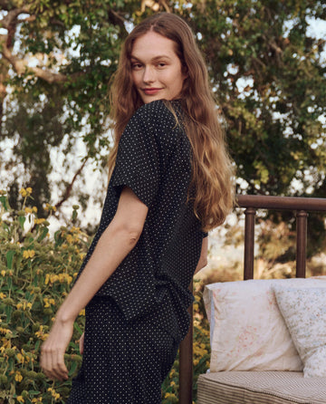 A person with long hair wearing THE GREAT. PS25 SLEEP's The Short Sleeve Pajama Shirt in Mini Black Polka Dot stands outdoors by a bed, surrounded by trees and yellow flowers.