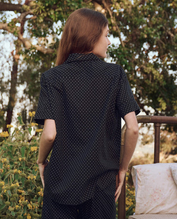 A person with long hair wearing THE GREAT. PS25 SLEEP's The Short Sleeve Pajama Shirt in Mini Black Polka Dot stands outdoors near a bed with floral sheets, surrounded by greenery and trees.