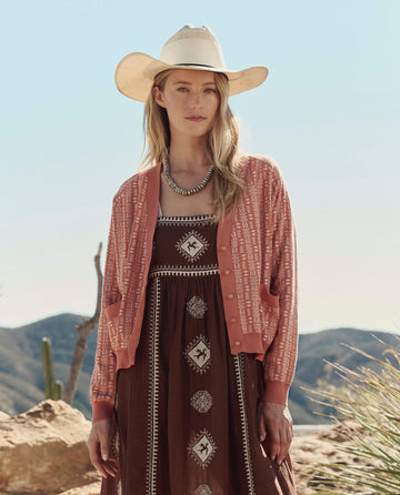 A woman wears THE GREAT. PF26 The Varsity Cardigan in Vintage Coral over a patterned brown dress and white cowboy hat, standing outdoors with desert mountains and cacti in the background.