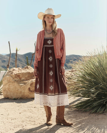A woman stands outdoors among desert plants and mountains, wearing THE GREAT. PF26’s The Varsity Cardigan in Vintage Coral with a cowboy hat, patterned dress, and brown boots.