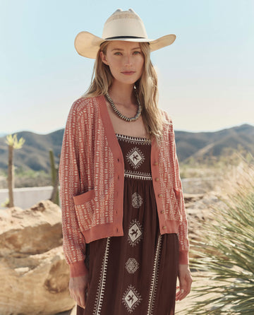 A woman wearing THE GREAT. PF26 The Varsity Cardigan in Vintage Coral and a patterned brown dress stands outdoors with mountains and desert plants in the background.