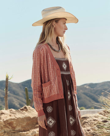 A woman in a cowboy hat wears THE GREAT. PF26 The Varsity Cardigan in Vintage Coral, standing outdoors before rocky terrain and distant mountains.