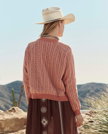A person wears THE GREAT. PF26 The Varsity Cardigan in Vintage Coral, paired with a wide-brimmed hat and long skirt, standing outdoors with mountains and cacti in the background.