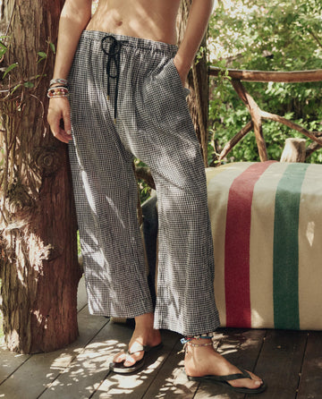 A person stands on a wooden deck wearing THE GREAT. SP26 SWIM's The Reef Pant in Black & Cream Check, paired with sandals and bracelets; a striped blanket appears in the background.
