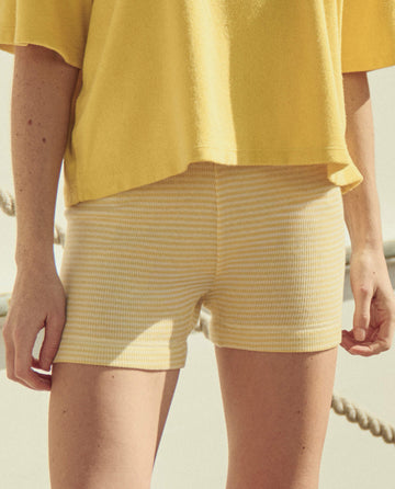 A person stands outdoors in THE GREAT. SU26 The Relay Short -- Soft Yellow Stripe, paired with a yellow top and a rope visible in the background.