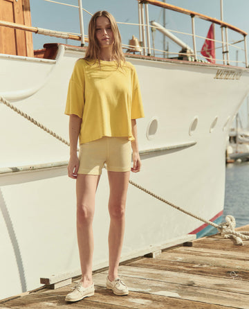 A woman stands on a wooden dock before a white yacht, wearing a yellow t-shirt, boat shoes, and THE GREAT. SU26 The Relay Short. -- Soft Yellow Stripe.
