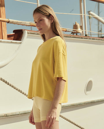 A woman stands on a boat deck in sunlight, wearing THE GREAT. SU26’s The Relay Short in Soft Yellow Stripe and a loose yellow top.