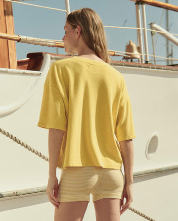 A woman with long hair stands outdoors facing a docked boat, wearing THE GREAT. SU26 The Micro Terry Track Crew in Margarita and light yellow shorts, capturing a relaxed track crew vibe.