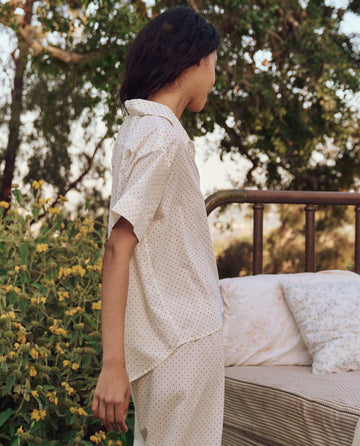 Standing near a metal bed frame with cushions, surrounded by greenery, is a person wearing THE GREAT. PS25 SLEEP's The Short Sleeve Pajama Shirt in Mini Cream Polka Dot.
