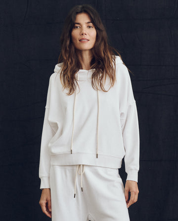 A woman with long dark hair is standing against a dark background, wearing The Teammate Hoodie in True White by THE GREAT. PS24 LOFTY KNITS and matching sweatpants, handmade in Los Angeles, looking at the camera.