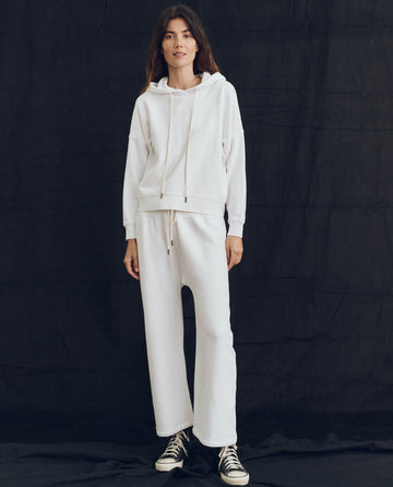 A person is standing against a dark background, wearing THE GREAT. PS24 LOFTY KNITS' The Teammate Hoodie in True White with matching white sweatpants and black sneakers.
