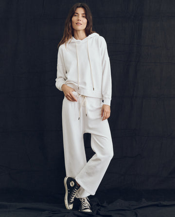 A person stands in front of a black backdrop, wearing "The Teammate Hoodie" in True White from THE GREAT. PS24 LOFTY KNITS, matched with white French Terry pants and paired with black sneakers.