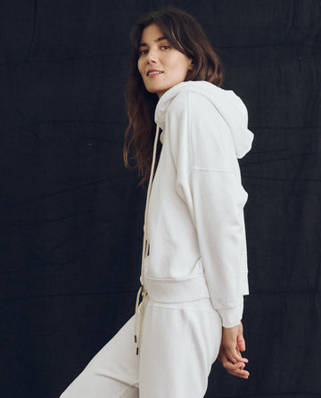 A person with long hair wears The Teammate Hoodie in True White from THE GREAT. PS24 LOFTY KNITS and matching French Terry sweatpants, standing against a dark background.