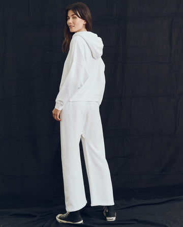 A person wearing THE GREAT. PS24 LOFTY KNITS' The Teammate Hoodie in True White and matching white pants stands against a black background, looking over their shoulder. They are also wearing black sneakers.
