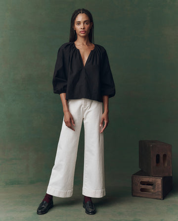 A woman faces forward in THE GREAT. F25 D2 The Chariot Top in black, paired with white wide-leg pants, dark socks, and black loafers, set against a green backdrop with two wooden boxes on the right.