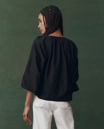 A person with braided hair wears THE GREAT. F25 D2's The Chariot Top in Black and white pants, standing against a green background and facing away from the camera.