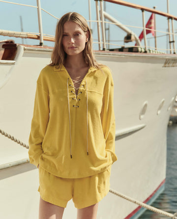 A woman stands before a yacht, wearing THE GREAT. SU26 Micro Terry Swing Short in Margarita and a yellow long-sleeve lace-up top.