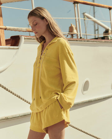 A woman in THE GREAT. SU26's Micro Terry Swing Short in Margarita stands beside a white boat, wearing a yellow long-sleeve top and looking down with her eyes closed.