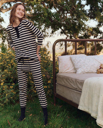 Wearing The Sleep Henley, Eventide Stripe by THE GREAT. PS25 SLEEP, a person stands outdoors beside a vintage metal bed adorned with floral bedding amidst lush greenery.