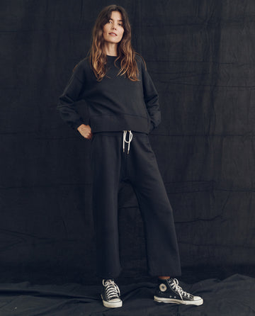 A woman stands against a dark backdrop wearing The League Sweatshirt in Almost Black from THE GREAT. PS24 LOFTY KNITS, along with black wide-legged pants featuring a white drawstring and black high-top sneakers. Her hands are in her pockets, and she has long, wavy hair. The ensemble exudes the charm of vintage athletic knits.