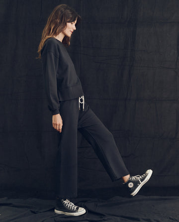 A woman stands sideways against a dark background, wearing The League Sweatshirt in Almost Black from THE GREAT. PS24 LOFTY KNITS, paired with cropped pants made from vintage athletic knits, and black sneakers with white laces and soles—handcrafted in Los Angeles.