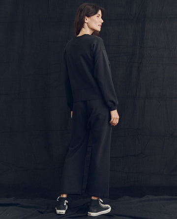 A person stands facing sideways, wearing The League Sweatshirt in Almost Black and wide-leg pants made from lofty loopback French Terry, paired with sneakers. The background is plain and dark.