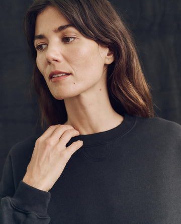 A person with long hair, wearing The League Sweatshirt in Almost Black by THE GREAT. PS24 LOFTY KNITS, handcrafted in Los Angeles, looks slightly to the side while touching the neckline of their garment against a dark background.